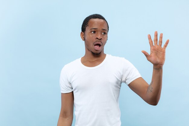 Cerca de medio cuerpo retrato de joven afroamericano con camisa blanca sobre fondo azul. Emociones humanas, expresión facial, publicidad, concepto de ventas. Conocer a alguien, saludar, invitar.