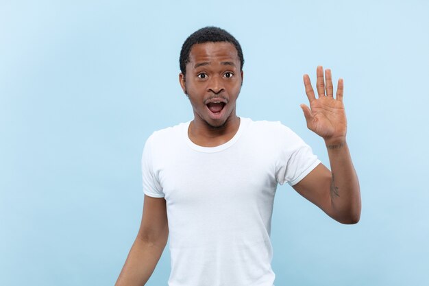 Cerca de medio cuerpo retrato de joven afroamericano con camisa blanca sobre fondo azul. Emociones humanas, expresión facial, publicidad, concepto de ventas. Conocer a alguien, saludar, invitar.