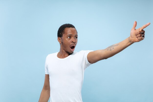 Cerca de medio cuerpo retrato de joven afroamericano con camisa blanca sobre fondo azul. Emociones humanas, expresión facial, publicidad, concepto de ventas. Señalando, eligiendo, asombrado. Copyspace.