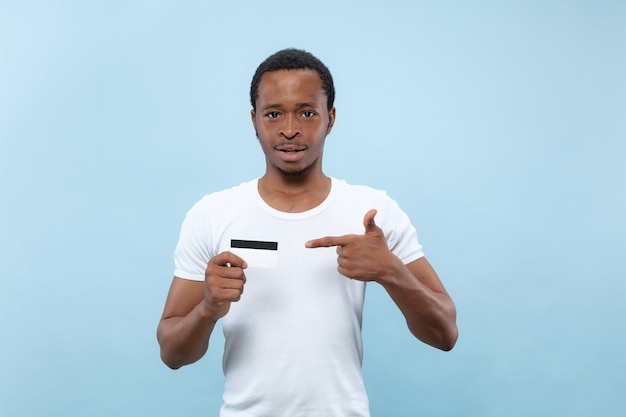 Cerca de medio cuerpo retrato de joven afroamericano con camisa blanca sobre fondo azul. Emociones humanas, expresión facial, publicidad, concepto de ventas. Señalando la tarjeta. Pagos, finanzas, factura.