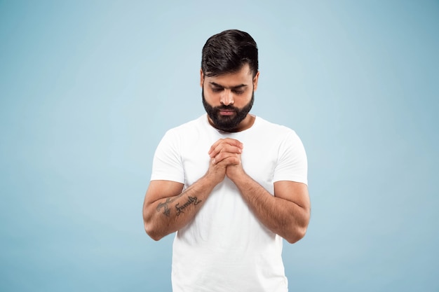 Cerca de medio cuerpo retrato de joven hindú con camisa blanca aislada en la pared azul. Las emociones humanas, la expresión facial, el concepto publicitario. Espacio negativo. De pie y rezando con los ojos cerrados.