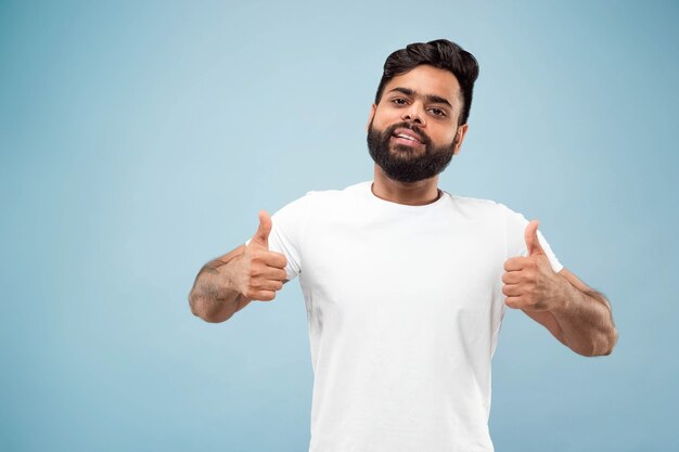 Cerca de medio cuerpo retrato de joven hindú con camisa blanca en la pared azul. Las emociones humanas, la expresión facial, el concepto publicitario. Espacio negativo. Mostrando el signo de OK, agradable, genial. Sonriente.