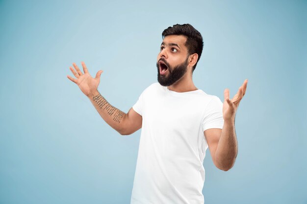 Cerca de medio cuerpo retrato de joven hindú con camisa blanca en la pared azul. Las emociones humanas, la expresión facial, el concepto publicitario. Espacio negativo. Sentimientos felices conmocionados, asombrados o locos.