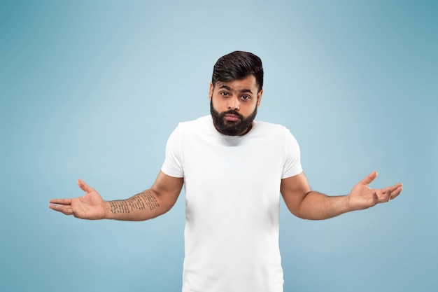 Cerca de medio cuerpo retrato de joven hindú con camisa blanca sobre fondo azul. Las emociones humanas, la expresión facial, el concepto publicitario. Espacio negativo. Confusión, desconocimiento, incertidumbre.