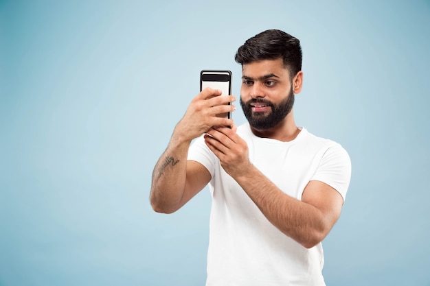 Cerca de medio cuerpo retrato de joven hindú con camisa blanca sobre fondo azul. Las emociones humanas, la expresión facial, el concepto publicitario. Espacio negativo. Hacer selfie o videoblog, vlog, chatear.