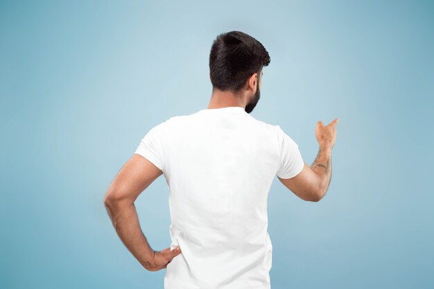 Cerca de medio cuerpo retrato de joven hindú con camisa blanca sobre fondo azul. Las emociones humanas, la expresión facial, el concepto publicitario. Espacio negativo. Mostrando barra vacía, señalando, eligiendo, invitando.