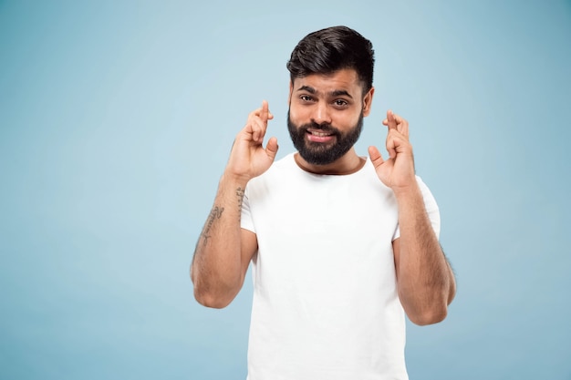 Cerca de medio cuerpo retrato de joven hindú con camisa blanca sobre fondo azul. Las emociones humanas, la expresión facial, el concepto publicitario. Espacio negativo. Mostrando el signo de la suerte, sonriendo.