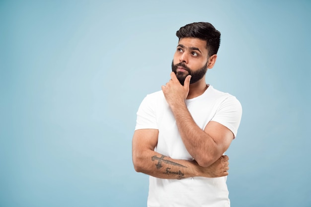 Foto gratuita cerca de medio cuerpo retrato de joven hindú con camisa blanca sobre fondo azul. las emociones humanas, la expresión facial, el concepto publicitario. espacio negativo. pensando mientras se lleva la mano a la barba. elegir.