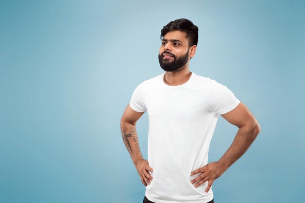 Cerca de medio cuerpo retrato de joven hindú con camisa blanca sobre fondo azul. Las emociones humanas, la expresión facial, el concepto publicitario. Espacio negativo. Posando, de pie y sonriendo, parece tranquilo.