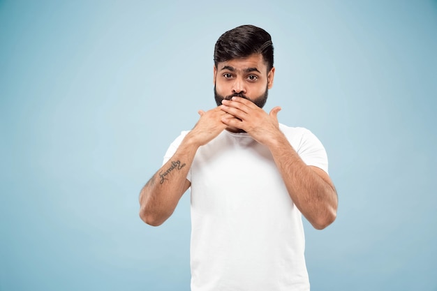 Cerca de medio cuerpo retrato de joven hindú con camisa blanca sobre fondo azul. Las emociones humanas, la expresión facial, las ventas, el concepto publicitario. Espacio negativo. Cubriéndose la cara con las manos.