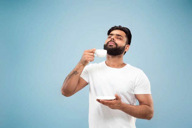 Cerca de medio cuerpo retrato de joven hindú con camisa blanca sobre fondo azul. Las emociones humanas, la expresión facial, las ventas, el concepto publicitario. Espacio negativo. Disfrutando de tomar café o té.