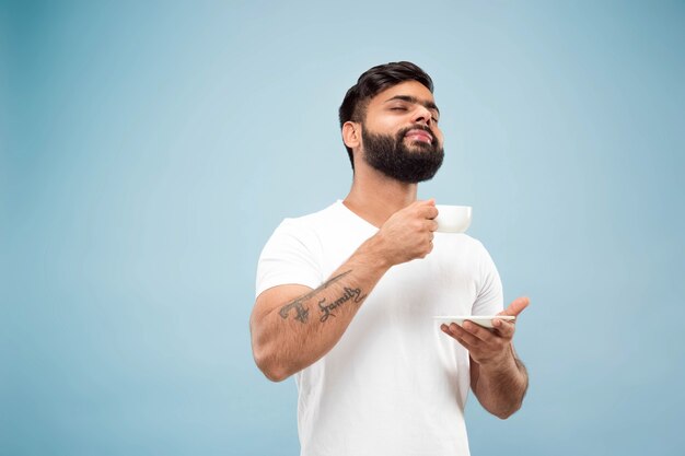 Cerca de medio cuerpo retrato de joven hindú con camisa blanca sobre fondo azul. Las emociones humanas, la expresión facial, las ventas, el concepto publicitario. Espacio negativo. Disfrutando de tomar café o té.