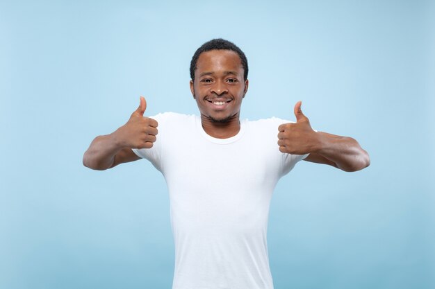 Cerca de medio cuerpo retrato de joven modelo masculino afroamericano en camisa blanca sobre fondo azul de estudio. Las emociones humanas, la expresión facial, el concepto publicitario. Mostrando el signo de OK. bonito.