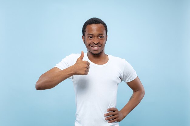 Cerca de medio cuerpo retrato de joven modelo masculino afroamericano en camisa blanca sobre fondo azul de estudio. Las emociones humanas, la expresión facial, el concepto publicitario. Mostrando el signo de OK. bonito.