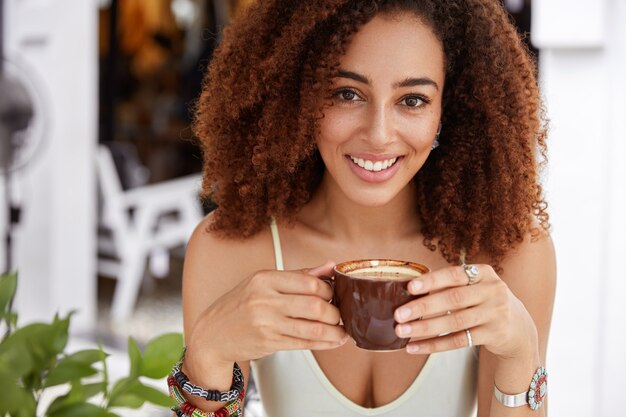 Cerca del modelo femenino afroamericano positivo sostiene una taza de café y mira felizmente a la cámara, recrea en un acogedor restaurante después de un duro día de trabajo