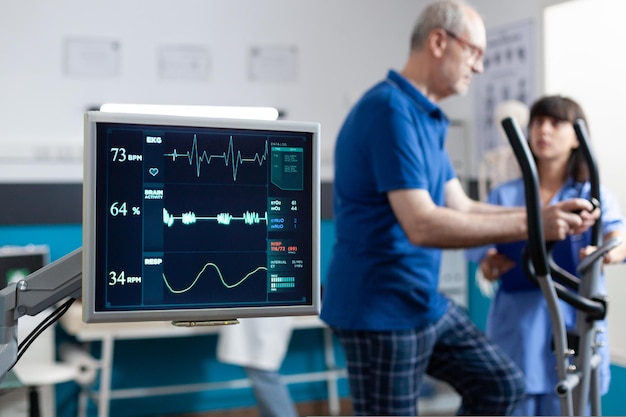 Cerca del monitor de frecuencia cardíaca que mide los latidos del corazón del paciente que hace gimnasia para la recuperación física. Hombre jubilado con equipamiento deportivo para hacer ejercicio de fisioterapia y fuerza.