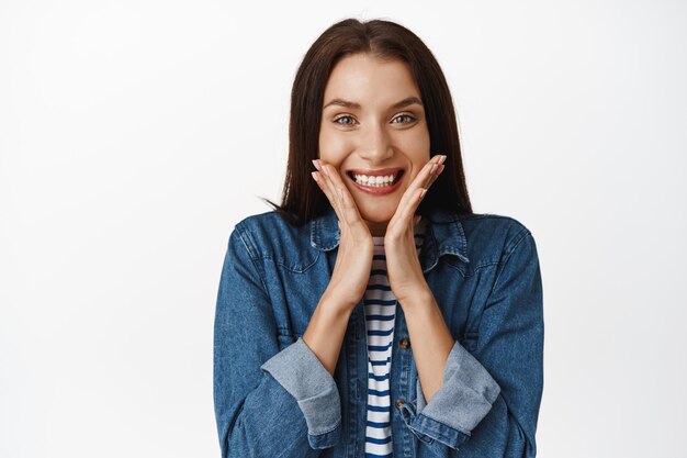 Cerca de una mujer adulta extremadamente feliz, dientes blancos sonrientes, tocando la cara natural limpia sin maquillaje y luciendo emocionada, de pie contra el fondo blanco.