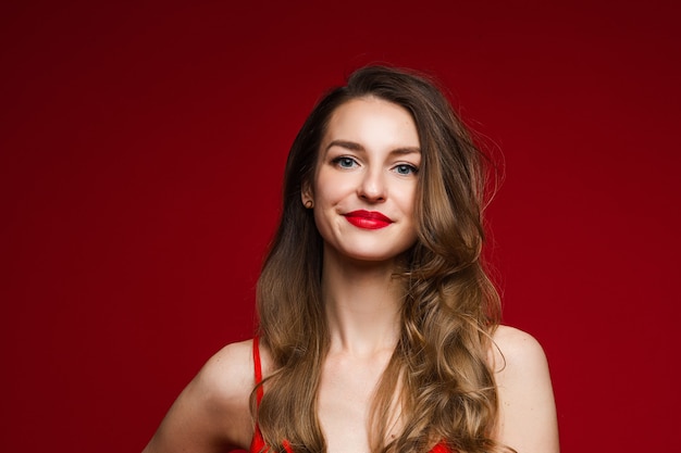 De cerca a una mujer adulta joven impresionante con largo cabello castaño ondulado sonriendo a la cámara con labios rojos regordetes. Aislado en rojo.