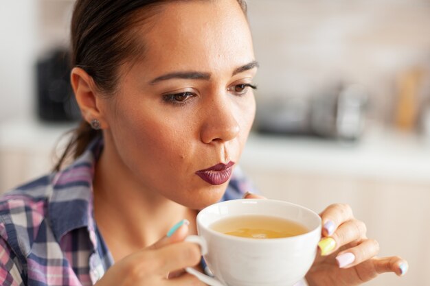 Cerca de la mujer en la cocina tratando de beber té verde caliente con hierbas aromáticas