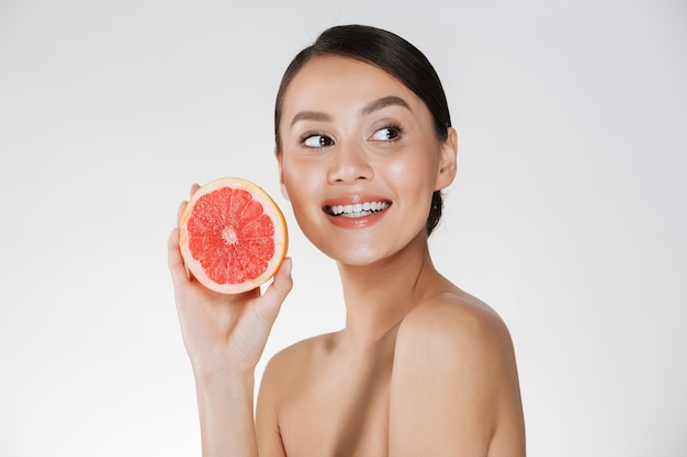 Cerca de la mujer contenta con una piel fresca y saludable con pomelo jugoso y mirando a un lado con una sonrisa, aislado en blanco