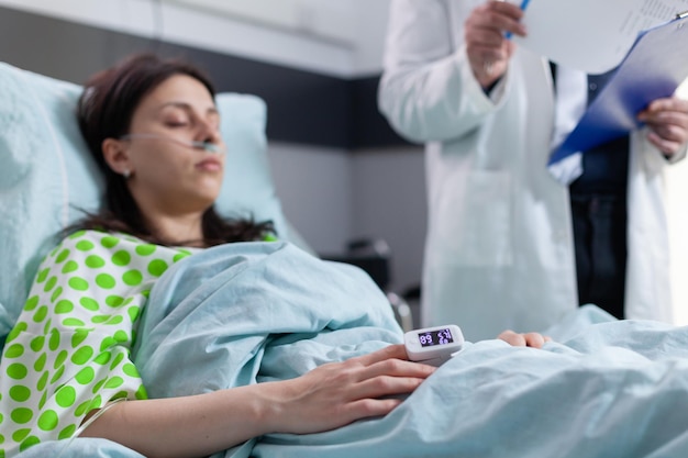 Cerca de una mujer enferma descansando en la cama con un monitor de frecuencia cardíaca en el dedo mientras el equipo médico revisa los latidos del corazón. Paciente con oxímetro esperando cardiograma de enfermedad en sala de hospital