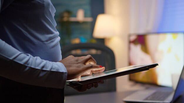 Cerca de mujer negra usando tableta, navegando, escribiendo en él, de pie en la sala de estar a altas horas de la noche tomando un descanso. Freelancer africano que usa la red inalámbrica de tecnología moderna haciendo horas extraordinarias