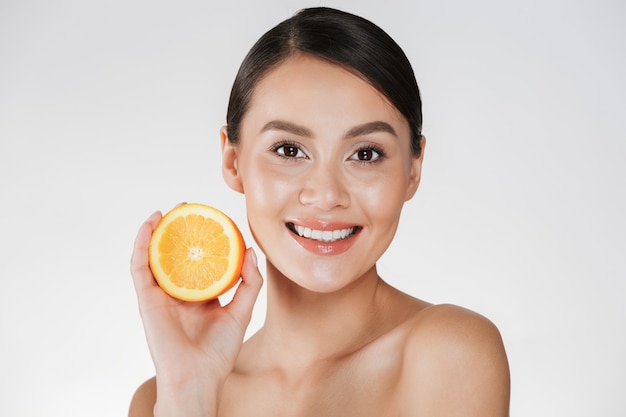 Cerca de una mujer satisfecha con una piel fresca y saludable con naranja jugosa y sonriente, aislado en blanco