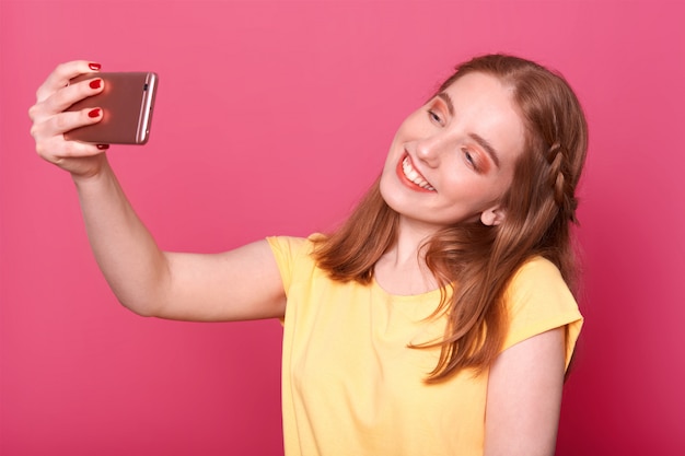 Cerca de mujeres jóvenes y elegantes haciendo selfie, usando su propio teléfono inteligente, vestida con una camiseta amarilla casual, tiene el pelo liso, quiere una nueva foto para las redes sociales