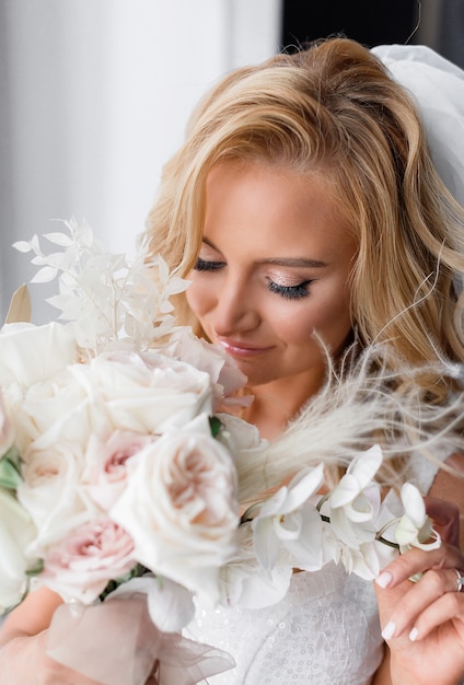 Cerca de la novia rubia con maquillaje natural, vestida con ropa de boda, sosteniendo un ramo de flores y disfrutando de su olor