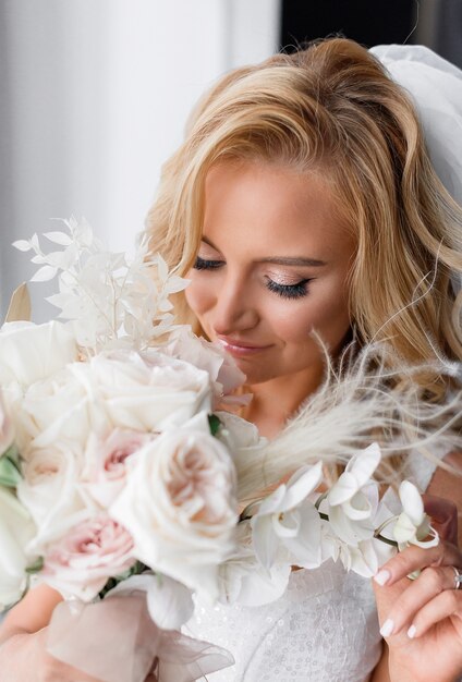 Cerca de la novia rubia con maquillaje natural, vestida con ropa de boda, sosteniendo un ramo de flores y disfrutando de su olor