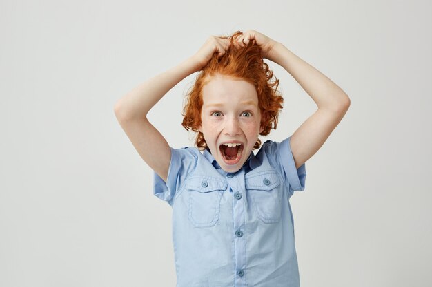 Cerca del pequeño y hermoso niño jengibre con pecas sosteniendo el pelo con las manos, gritando