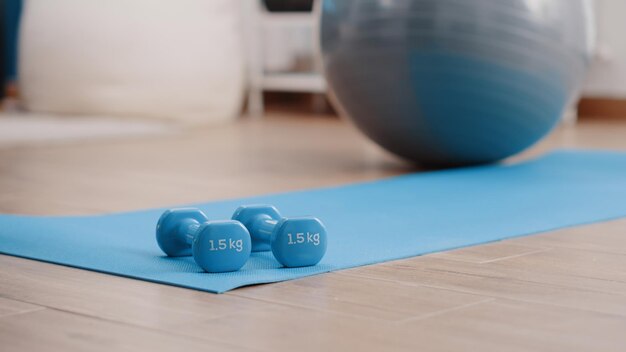 Cerca de pesas azules en la estera de yoga del piso para entrenar los músculos y hacer ejercicio físico en casa. Espacio vacío con levantamiento de pesas utilizado para trabajar en brazos. Equipo deportivo y de entrenamiento.