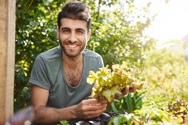 Cerca de retrato de un hermoso agricultor caucásico barbudo de piel oscura sonriendo, trabajando en el jardín, recolecta hojas de lechuga, preparándose para la reunión nocturna con amigos en su casa
