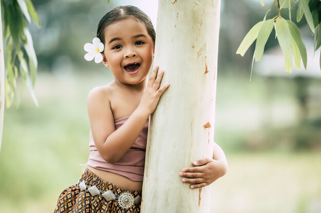 De cerca, Retrato de niña en traje tradicional tailandés y poner flor blanca en su oreja, Párese y abrace el tronco del árbol, ríe, copie el espacio