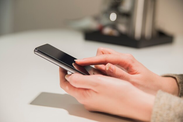 Cerca de smartphone en la mano. Ciérrese para arriba de una mano de la mujer que sostiene un smartphone y texting. Manos de womans escribiendo en smartphone sobre mesa de madera marrón