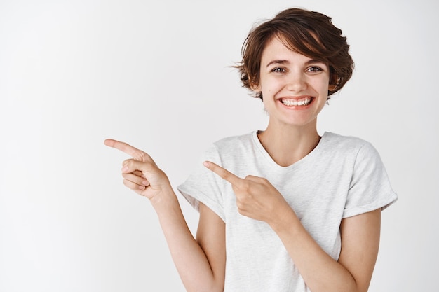 Cerca de sonriente mujer caucásica con peinado corto y sin maquillaje apuntando a la izquierda en el espacio vacío, pared blanca