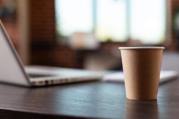 Cerca de la taza de café desechable y la computadora portátil en el escritorio de la oficina en el espacio de inicio. Bebida caliente en la mesa en el lugar de trabajo vacío. Beber en vaso de plástico sobre muebles y computadora.