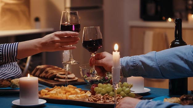 Cerca de tintineo de copas de vino tinto durante una cena romántica. Feliz pareja joven alegre cenando juntos en la acogedora cocina, disfrutando de la comida, celebrando el brindis romántico aniversario