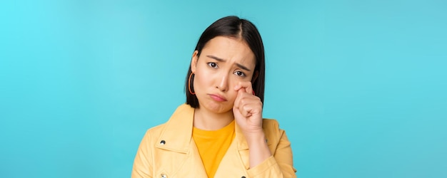 Cerca de una triste chica asiática llorando limpiando lágrimas y enfurruñada luciendo molesta y sombría posando contra b