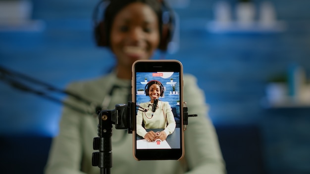 Cerca de vlog de grabación de smartphone de influencer africano en estudio casero con smartphone. Hablando durante la transmisión en vivo, bloguero discutiendo en podcast usando audífonos y micrófono profesional