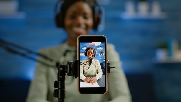 Cerca de vlog de grabación de smartphone de influencer africano en estudio casero con smartphone. Hablando durante la transmisión en vivo, bloguero discutiendo en podcast usando audífonos y micrófono profesional