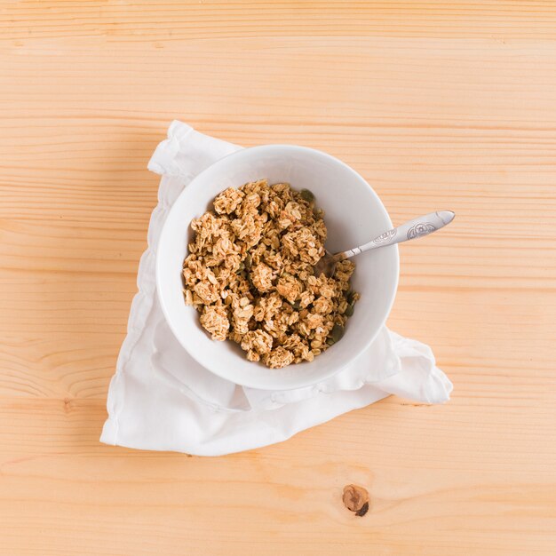 Cereal y cuchara de desayuno del granola de la avena en el cuenco blanco sobre la servilleta blanca en el escritorio de madera