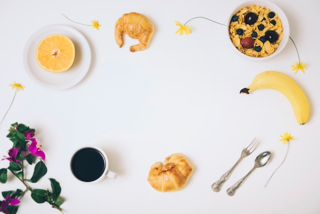 Cereales de maíz; plátano; croissants Copa de naranja y café a la mitad con flor de buganvilla sobre fondo blanco