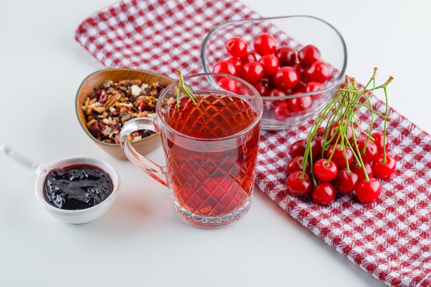 Cereza en un tazón con té, mermelada, hierbas secas vista de ángulo alto en blanco y papel de cocina