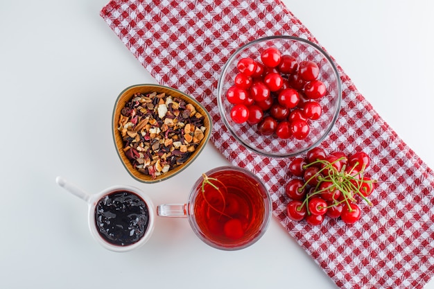 Cereza con té, mermelada, hierbas secas en un tazón blanco y papel de cocina, plano.