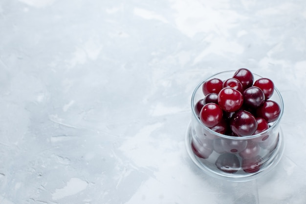 Cerezas agrias frescas dentro de un pequeño vaso de vidrio sobre un escritorio blanco claro, fruta agria vitamina vitamina foto