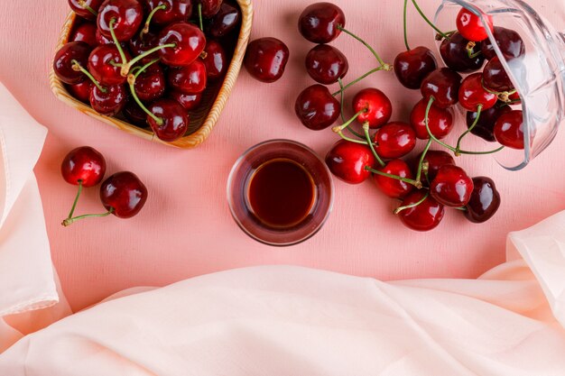 Cerezas en florero y plato con vaso de té vista superior en superficie rosa y textil