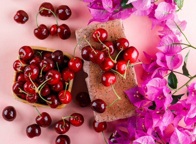 Cerezas con flores, ladrillo en una placa de madera sobre superficie rosa