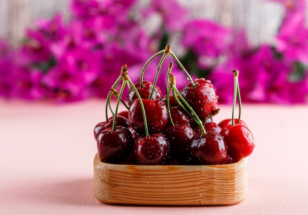 Foto gratuita cerezas con flores en un plato de madera sobre superficie rosa, vista lateral