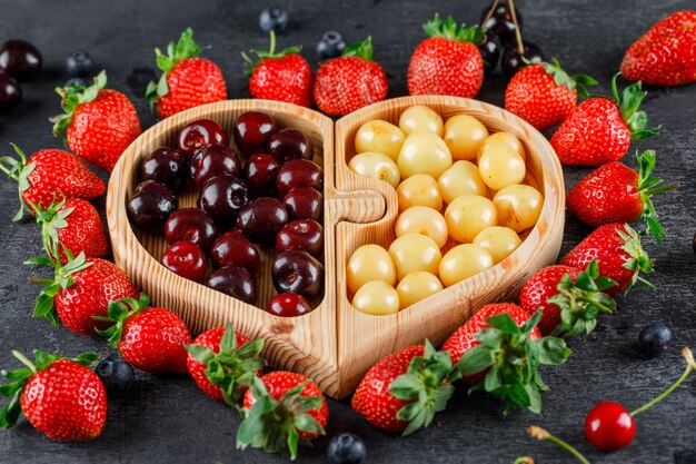 Cerezas con fresas, arándanos en una placa de madera sobre la superficie gris, vista de ángulo alto.